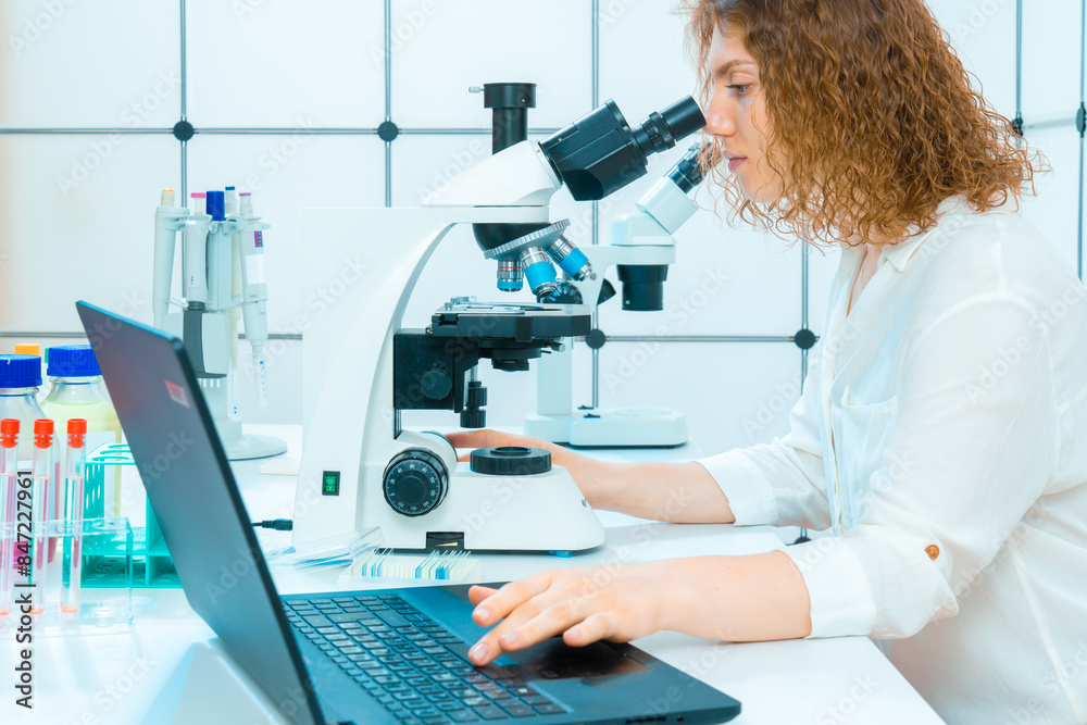 Poster female laboratory assistant in a science laboratory with a microscope and laptop