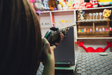 A young girl aiming a rifle at a target in a shooting game booth filled with toy prizes, an over-the-shoulder shot.