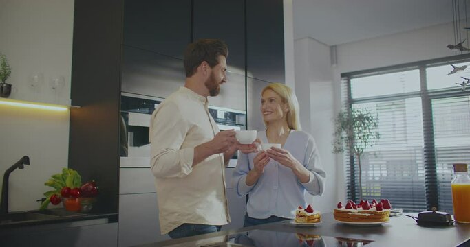 Caucasian male walking up to his wife. People standing in front of countertop in middle of large kitchen. Man giving cup of hot drink to woman. Enjoying their time together while eating delicious cake