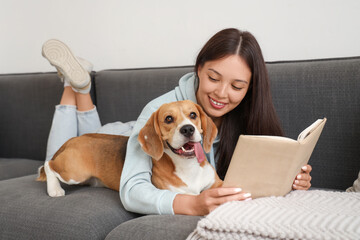 Young woman reading book and lying with adorable Beagle dog on sofa in living room