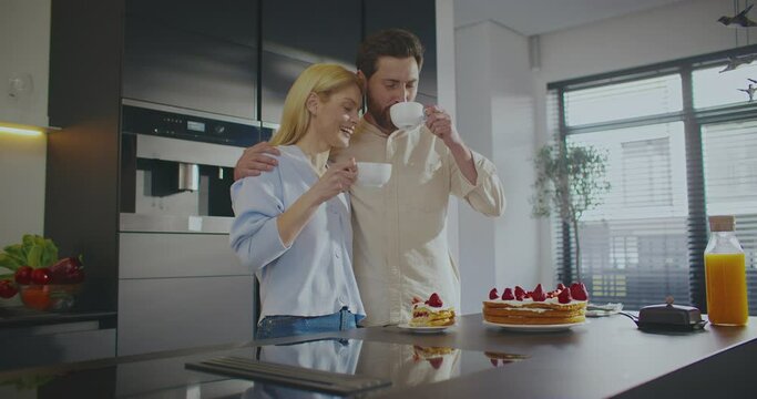 Lovely Caucasian couple standing together in middle of modern kitchen. Husband hugging his beautiful wife. Enjoying their morning drinks. Hot coffee or tea. Eating cake while talking to each other.
