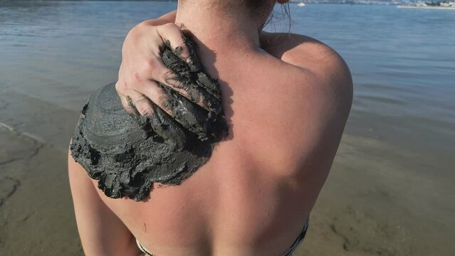 Woman  applying healing mud on back and shoulder with her hand, thalassotherapy and balneotherapy by black peliod mud  at the beach Soline on the Island of Krk, Croatia