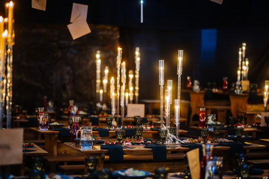 Dobele, Latvia - June 7, 2024 - Elegant dining setup with tall glass candle holders, floating envelopes, wooden tables, glassware, and plates, creating a warm and festive atmosphere. Copy space.