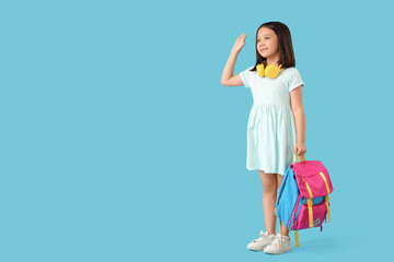 Cute little happy Asian girl with backpack and headphones waving hand on blue background. End of school year