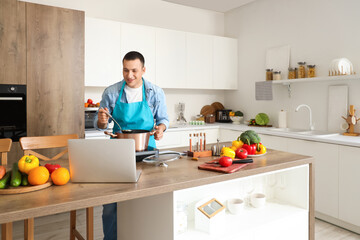 Young man with laptop cooking soup in kitchen