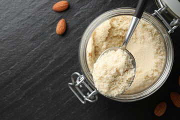 Spoon with fresh almond flour above jar and nuts on dark textured table, top view. Space for text