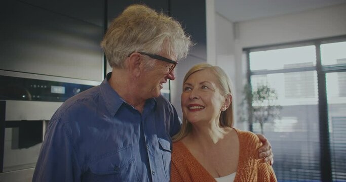 Camera focus on loving Caucasian couple. Husband hugging beautiful wife in middle of kitchen. In background oven and microwave built into wall. Large windows with sun shining bright outside.