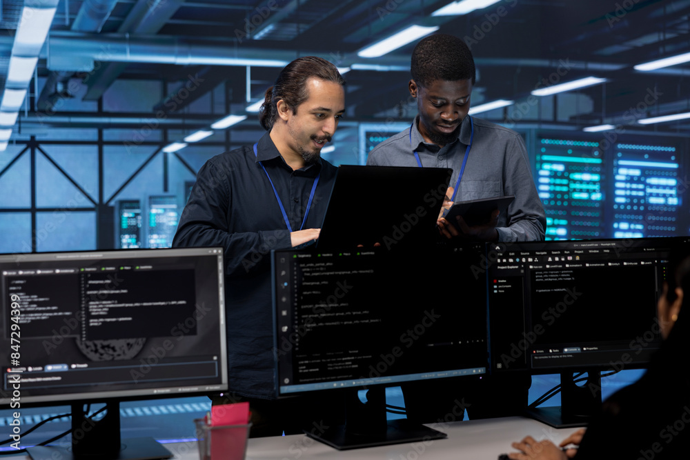 Poster coworkers in data center using laptop and tablet to find firewall misconfigurations affecting server