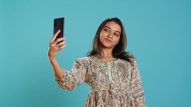 Cheerful indian woman using smartphone to take selfies and post them on social media. Happy person taking photos using phone selfie camera, isolated over studio background, camera A