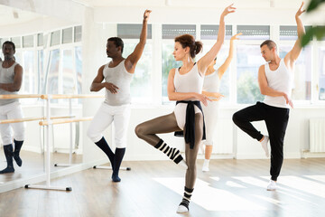 Graceful woman confidently leading ballet class with adult male and female amateur dancers following her movements in spacious sunlit dance studio