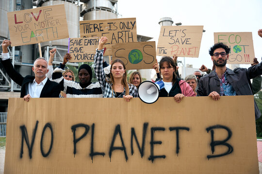 Group of people of different ages and nationalities with arms raised in protest against factories and pollution. Pro-earth demonstration with climate change banners looking at camera with serious face