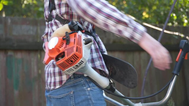 Man with electric lawnmower, lawn mowing. Gardener trimming garden. Suburb, village. Young caucasian male farmer gardener standing in yard with petrol trimmer. Brush cutter cutting weeds on farm.