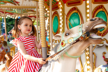 Adorable little brunette girl in summer red-white dress at amusement park having a ride on the...