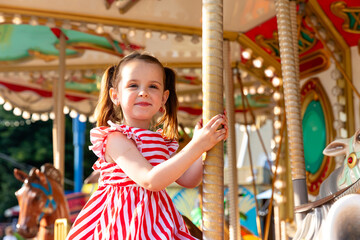 Adorable little brunette girl in summer red-white dress at amusement park having a ride on the merry-go-round. Child girl has fun outdoor on sunny summer day. Entertainment concept © Irina Mikhailichenko