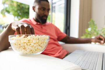 African American young man relaxing at home, reaching for popcorn while using laptop, at home