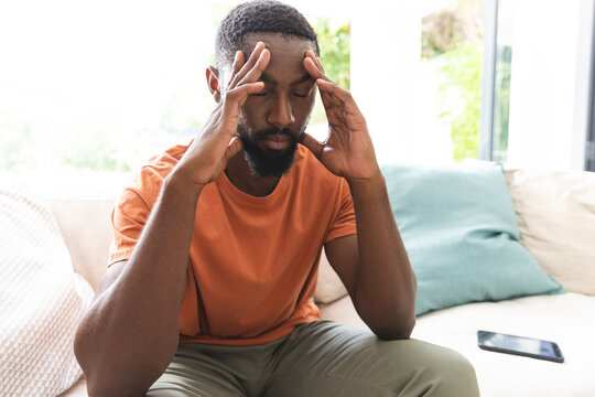 African American young man sits on couch, looking stressed with hands on temples