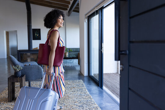 Biracial young woman with curly hair carrying suitcase and red bag, vacation home