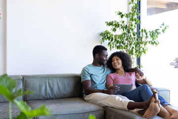 A diverse couple relaxing on couch, woman holding a tablet and smiling at the man, copy space