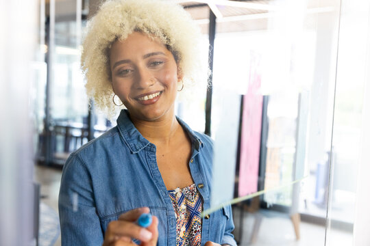 Young biracial woman writing on glass board, smiling at camera in business office 