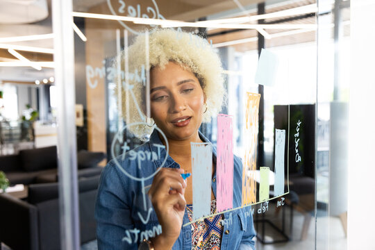 Young biracial woman drawing colorful bar graph on glass board in modern business office