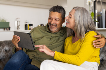 A biracial mature couple sits on the couch, smiling and looking at a tablet, at home