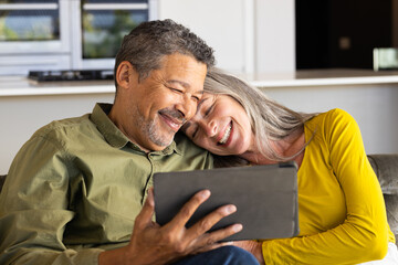 A biracial mature couple is smiling while looking at tablet together, at home