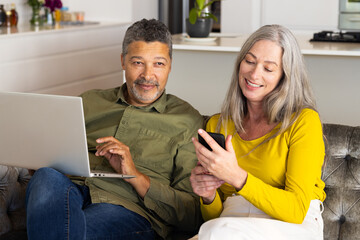 Biracial mature couple relaxing at home, interacting with laptop and smartphone, at home