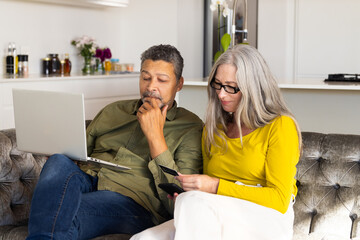 A biracial mature couple sits on a couch: the man uses a laptop while the woman checks her smartphon