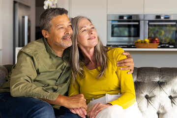 A biracial mature couple is sitting on couch, smiling and looking content, at home