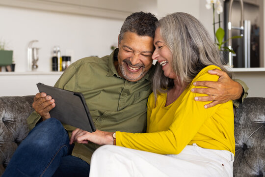 A biracial mature couple is sitting on couch, smiling and looking at a tablet, at home - Powered by Adobe