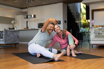 A biracial mature couple is sitting on yoga mats, smiling and taking selfie, at home