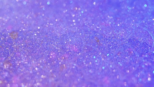 Shiny fluid. Glitter paint flow. Defocused purple color shimmering sequin particles flicker in translucent water liquid emulsion spill motion abstract art background with bokeh lights.