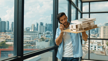 Smart caucasian engineer standing and holding house model while phone calling to manager....