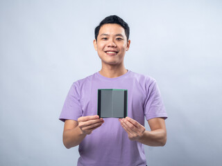 Smiling young man in a lavender shirt, holding a small notebook with a gray cover towards the camera against a plain light gray background