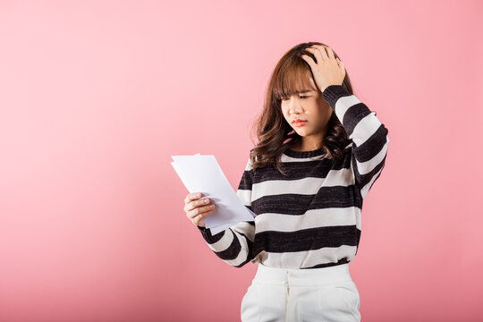 Portrait of beautiful Asian young woman sad tired strain holding paper calculating bills on hand, female person problem she hold worried over bill, studio shot isolated on pink background