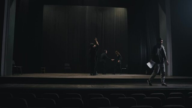 Confident expressive stage director in a gray T-shirt and scarf leaves the stage while his actors in black suits rehearse their roles and script on stage in the theater