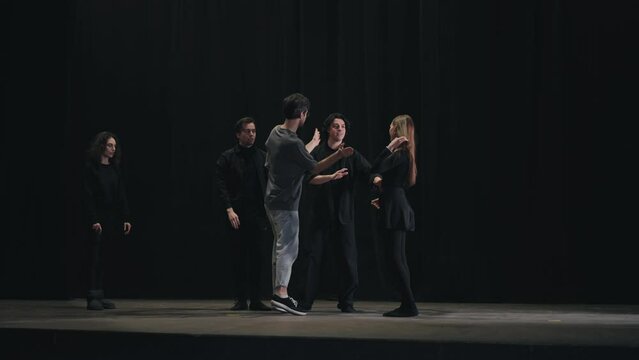 A confident stage director a brunette man in a gray T-shirt tells the actors how to correctly play the beats on stage during their rehearsal for a performance in the theater. A young girl actor slaps