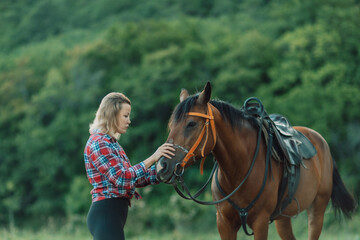 Obraz premium Happy blonde with horse in forest. Woman and a horse walking through the field during the day. Dressed in a plaid shirt and black leggings.