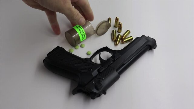 person picking up a black gun firearm with empty pill medicine bottle with a caution drowsiness sign and pills next to the gun firearm surrounded by some 9mm bullets