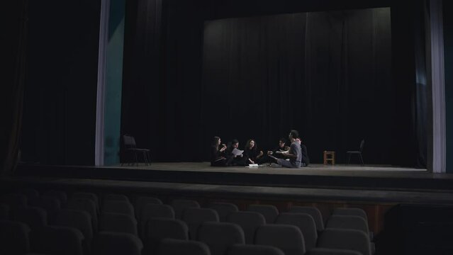 A group of young actors in black uniforms sit on stage together with their stage director communicate and analyze the script while preparing for a performance in a theater with a black curtain