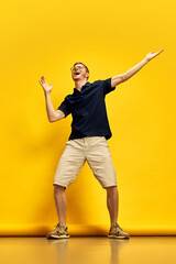 Full-length image of young man in dark blue polo shirt and beige shorts dancing against bright yellow background. Concept of human emotions, facial expression, lifestyle, fun