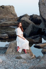 Serene woman in white dress sitting by ocean with red shoes on a rock gazes out at sea