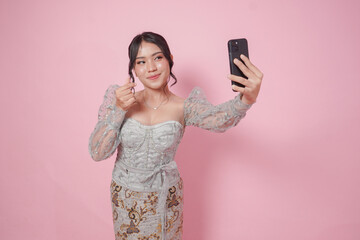 Cute Indonesian woman in modern kebaya posing for a selfie while making finger heart gesture, isolated by pink background.