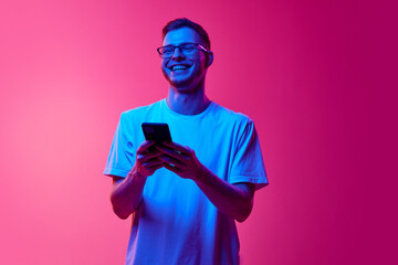 Laughing young man in glasses and casual clothes using mobile phone for communication against pink...