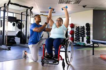 Quadriplegic biracial woman in wheelchair exercising with dumbbells, assisted by trainer they are in