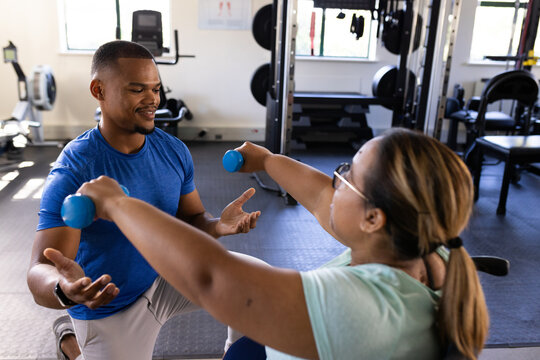 Quadriplegic biracial woman in wheelchair exercising with dumbbells, assisted by trainer