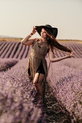 A woman is standing in a field of purple flowers, wearing a black dress and a black hat.