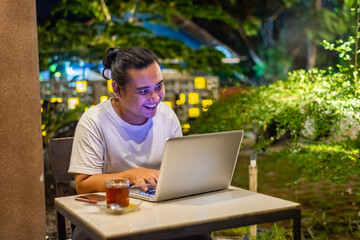 Young Asian man, digital nomad working remotely from cafe, using laptop and smartphone. young Asian men are hanging out in a cafe with a nature theme at night