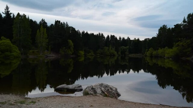 A serene view of Pinders Pond near the town of Roxburgh in New Zealand. The calm water reflects the surrounding trees and sky, creating a peaceful and picturesque scene