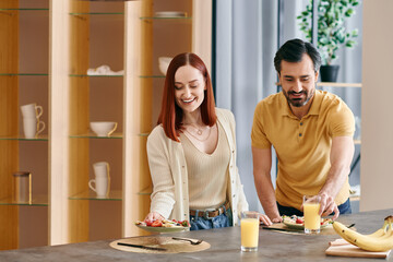 A redhead woman and bearded man prepare ingredients and cook together in a modern apartment kitchen.